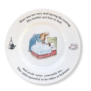 Peut inclure: Assiette en céramique blanche avec une illustration enfantine d'un lapin au lit, recevant du thé à la camomille. Une théière et une tasse à thé sont également représentées. Le texte dit : "Peter n'allait pas très bien pendant la soirée. Sa mère l'a mis au lit, et a fait du thé à la camomille : Une cuillère à soupe à prendre au coucher."