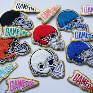 May include: Embroidered football helmet patches in black, red, orange, white, and blue. Each patch has a gold glitter border and a pennant with the text "GAMEDAY". The patches are designed for sports fans.