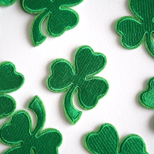 May include: A collection of green shamrock patches with embroidered detailing, scattered on a white background. The shamrocks are a vivid green, with a textured appearance. Perfect for St. Patrick's Day themed crafts and decorations.