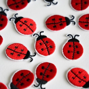 Könnte beinhalten: Eine Ansammlung gestickter Marienkäfer-Patches. Jeder Patch zeigt einen roten Körper mit schwarzen Markierungen und einem weißen Rand. Die Marienkäfer haben schwarze Fühler und liegen auf einer weißen Oberfläche.