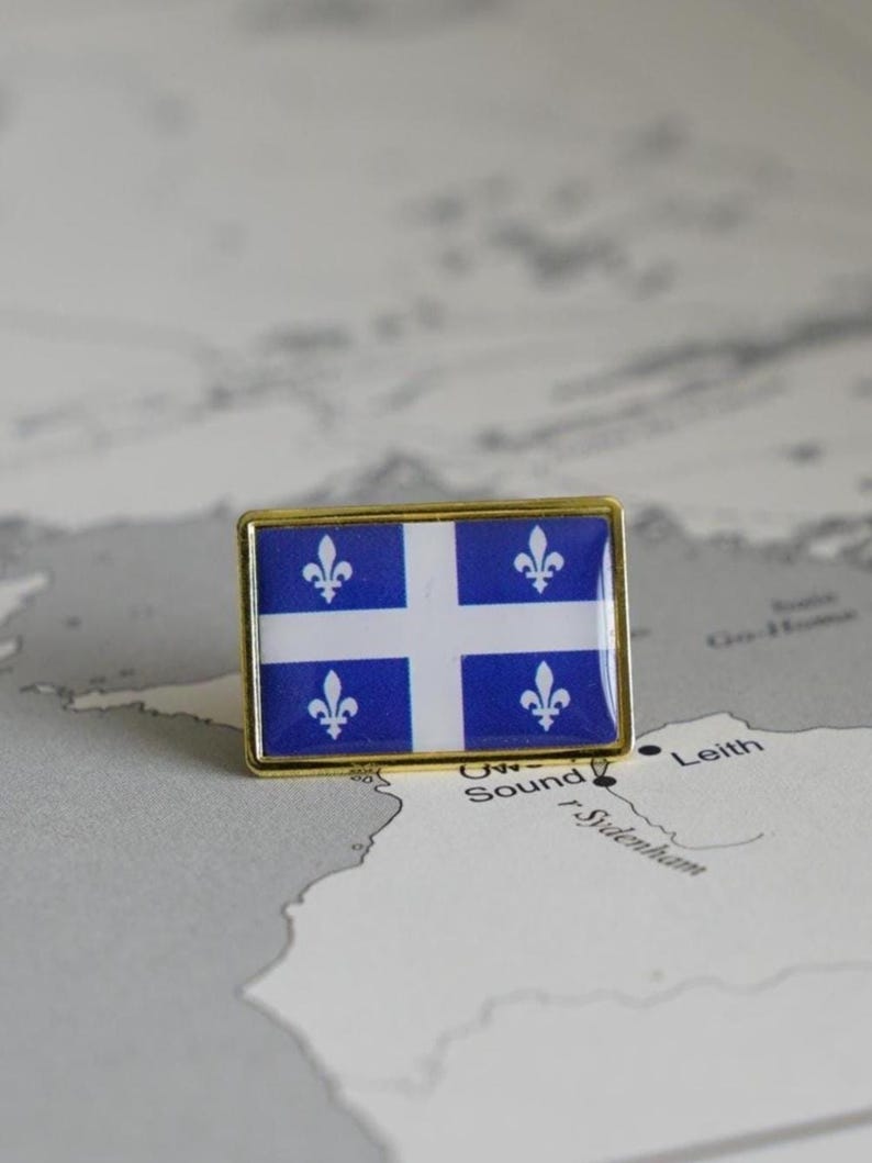 May include: A gold-colored pin with the flag of Quebec. The flag is blue with a white cross and four white fleurs-de-lis.