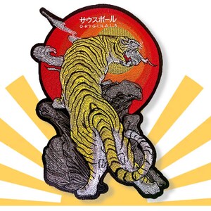 May include: Embroidered patch depicting a yellow and black tiger against a red sun. The tiger is positioned on a rock, with the text "サウスポール ORIGINALS" above the sun. The patch is set against a white backdrop with yellow sun rays.