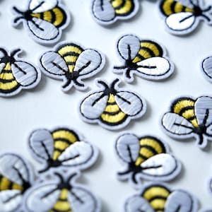 Könnte beinhalten: Nahaufnahme von mehreren gestickten Bienenaufnähern. Jeder Aufnäher hat einen gelb-schwarz gestreiften Körper, graue Flügel und eine weiße Umrandung. Die Aufnäher sind auf einer weißen Oberfläche verstreut und erzeugen ein verspieltes und skurriles Design.