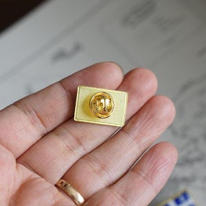 May include: The back of a gold-colored pin with a safety pin closure. The pin is rectangular and has a smooth, shiny surface.