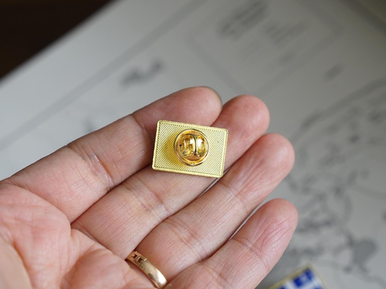 May include: The back of a gold-colored pin with a safety pin closure. The pin is rectangular and has a smooth, shiny surface.