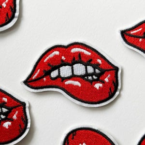 May include: Red embroidered lips with white teeth, a playful and bold design for a patch or iron-on.