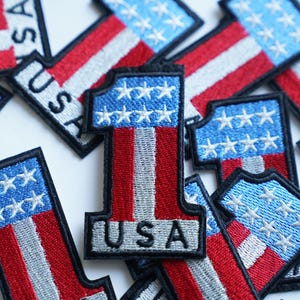 Può includere: Una collezione di toppe ricamate con il numero "1" con un design di bandiera americana e la parola "USA".