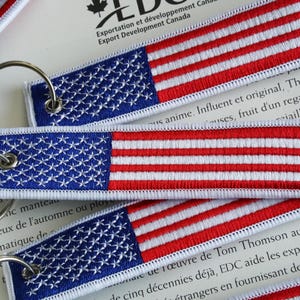 Puede incluir: Llaveros bordados con el diseño de la bandera estadounidense. Cada llavero tiene un campo azul con estrellas blancas y rayas rojas y blancas. Los llaveros están sujetos a anillos de metal plateado.