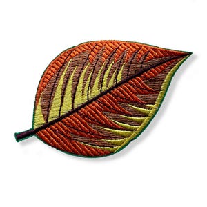 May include: Embroidered leaf patch in shades of orange, brown, and yellow, with a dark green border. The leaf has a detailed, textured design, and a dark stem. The patch is likely for clothing or accessories.