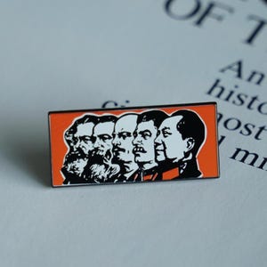 Może przedstawiać: Prostokątna emaliowana przypinka z pomarańczowym tłem przedstawia czarno-białe portrety pięciu postaci historycznych. Przypinka ma czarną obwódkę i jest umieszczona na rozmytym tle drukowanego tekstu.