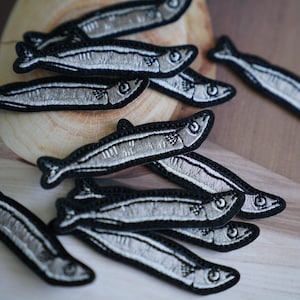 Könnte beinhalten: Eine Sammlung von gestickten Bügelbildern mit silbernen Fischen auf schwarzem Hintergrund. Die Patches sind auf einer Holzoberfläche angeordnet.