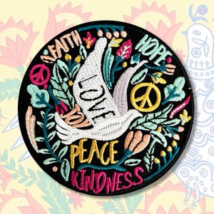 Op de afbeelding: Gestikte patch met een witte duif die over een kleurrijk bloemenmotief vliegt. De woorden "Geloof, Hoop, Liefde, Vrede, Goedheid" zijn rond de duif gestikt.