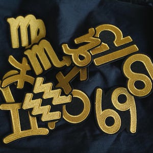 Op de afbeelding: Een verzameling van twaalf gouden sterrenbeeld-patches, elk met een zwarte omtrek. De patches zijn in willekeurige volgorde op een donkerblauwe stoffen achtergrond gerangschikt.