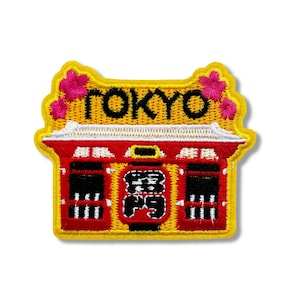 May include: An embroidered patch featuring a traditional Japanese building with a white roof and red doors. The building is decorated with pink flowers. The patch is red and yellow with the word "TOKYO" in black lettering.