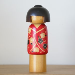 Peut inclure: Poupée Kokeshi japonaise en bois, avec une tête noire arrondie et un kimono rouge orné de motifs floraux dorés et noirs. La poupée a un corps cylindrique et une base en bois naturel.