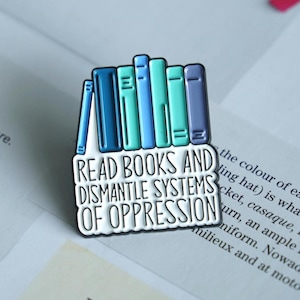 May include: An enamel pin with a design of stacked books in blue and green hues. The pin includes the text "READ BOOKS AND DISMANTLE SYSTEMS OF OPPRESSION" in white lettering, set against a black border.