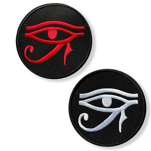 May include: Two circular patches with an embroidered Eye of Horus design. One patch features a red eye on a black background, the other a white eye on black. These patches are likely for clothing or accessories.