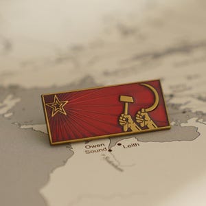 May include: A red enamel pin with a gold border depicting a hammer and sickle with a red star in the background.