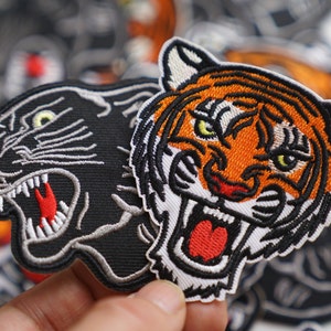 Può includere: Due toppe ricamate da stirare, una di una pantera nera e una di una tigre arancione e nera, entrambe con la bocca aperta e denti affilati.