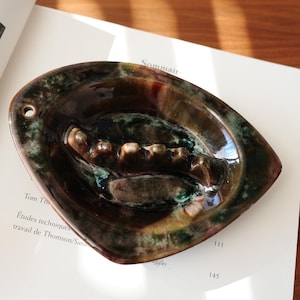 May include: A glossy, teardrop-shaped ceramic ashtray featuring a deep brown and forest green mottled glaze. The centre includes a raised, segmented ridge for resting items, and a small circular hole is positioned on the left side edge.