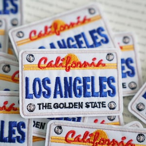 Pode incluir: Um conjunto de patches bordados brancos com um design de placa de matrícula da Califórnia. Os patches apresentam o texto "California" e "Los Angeles" em letras vermelhas e azuis, com um gráfico de sol amarelo. O texto "The Golden State" também está incluído em letras pretas.
