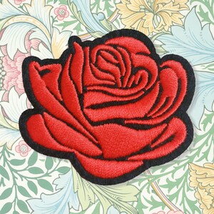 May include: Embroidered red rose patch with a black border. The rose features detailed textured stitching, showing the petals and centre. The patch is set against a floral patterned background, giving a vintage aesthetic.
