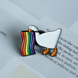 Peut inclure: Épingle en émail représentant une oie blanche avec des pattes et un bec orange, tenant un drapeau arc-en-ciel. Le pin's a un contour noir et est posé sur un fond de texte imprimé.