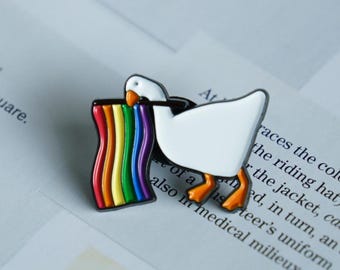 May include: A white goose enamel pin with orange feet and beak, clutching a rainbow flag. The pin features a black outline and is set against a backdrop of printed text.