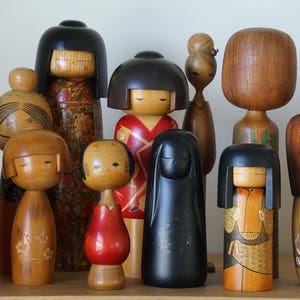 May include: A collection of wooden Kokeshi dolls of varying sizes and designs. The dolls feature painted faces, cylindrical bodies, and some have decorative clothing patterns in red, orange, and black. The dolls are displayed on a wooden surface.