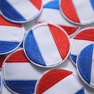 Könnte beinhalten: Nahaufnahme mehrerer runder Stoffaufnäher. Jeder Aufnäher zeigt ein Design mit roten, weißen und blauen Farben, das einer Flagge ähnelt. Die Aufnäher sind weiß mit einem genähten Rand, und die Farben sind in einem vertikalen oder diagonalen Muster angeordnet.