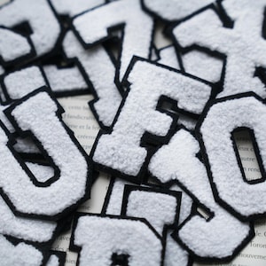 Może przedstawiać: Białe filcowe litery z czarnym obramowaniem rozrzucone na stronie książki. Litery w stylu uniwersyteckim mają puszystą fakturę. Litery są w różnych rozmiarach i orientacjach, tworząc efekt dekoracyjny. Litery wydają się być naszywkami lub aplikacjami.