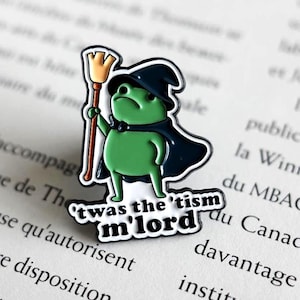 Peut inclure: Une broche en émail représentant une grenouille verte portant un chapeau et une cape de sorcier bleu foncé, tenant un balai marron. Sous la grenouille, une bannière blanche avec le texte noir : 'twas the 'tism m'lord. Finition brillante.
