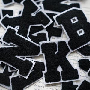 Pode incluir: Um conjunto de patches de letras de feltro preto com contornos brancos. As letras são de vários tamanhos e estilos.