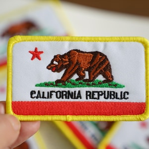 May include: A yellow-bordered patch with the California state flag. The flag features a red star, a brown bear walking on green grass, and the text "CALIFORNIA REPUBLIC".