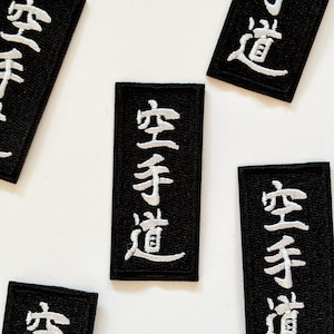 Könnte beinhalten: Auf weißem Hintergrund sind mehrere schwarze, rechteckige Aufnäher mit weißen japanischen Schriftzeichen verteilt. Die gestickten Aufnäher zeigen den Text "空手道". Diese Aufnäher sind wahrscheinlich für Kampfsportarten gedacht.
