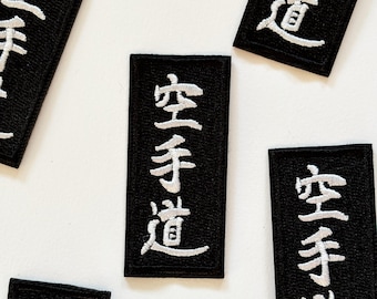 May include: Black rectangular patches with white Japanese characters are scattered across a white surface. The patches appear to be embroidered, with the text "空手道" prominently displayed. The patches are likely for martial arts enthusiasts.