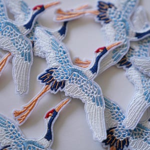 May include: A collection of embroidered iron-on patches featuring blue and white cranes with red beaks. The patches are perfect for adding a touch of nature and whimsy to clothing, bags, or other fabric items.