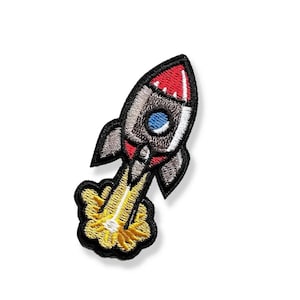 May include: Embroidered patch featuring a rocket ship design. The rocket is mainly grey with red and white details, a blue porthole, and yellow flames. The patch has a black border and is set against a white backdrop.