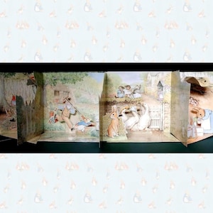 May include: A multi-paneled pop-up book featuring detailed watercolour illustrations of Peter Rabbit and other characters in a garden setting. The scenes depict rabbits, ducks, and a kitchen interior with soft, muted earth tones and green foliage.