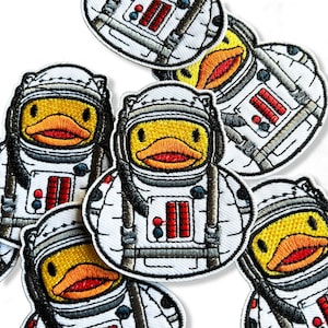 Op de afbeelding: Een set geborduurde strijkpatches met gele rubberen eendjes die verkleed zijn als astronauten. De patches zijn wit met zwarte en grijze details. Elke patch heeft een rood en wit bedieningspaneel op de borst.