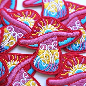 May include: A close-up of several embroidered iron-on patches featuring colourful psychedelic mushrooms with a sun and wave design. The patches are red, pink, yellow, blue, and white.
