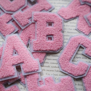 Könnte beinhalten: Nahaufnahme eines Stapels rosa Filzbuchstaben. Die Buchstaben sind in verschiedenen Größen und alle in einem hellen Rosaton. Die Buchstaben befinden sich auf einer weißen Oberfläche.