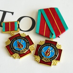 May include: Two gold-colored medals with red and blue accents, featuring a central emblem and Cyrillic text. Each medal has a red and green striped ribbon. The medals are likely commemorative or service awards.
