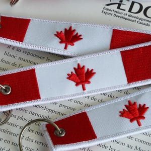 May include: Embroidered keyrings featuring the Canadian flag design. Each keyring has a red maple leaf on a white background, with red bars on each end. The keyrings are attached to silver metal rings.