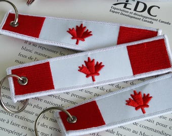 May include: Embroidered keychains featuring the Canadian flag design. Each keychain has a red maple leaf on a white background, with red bars on each end. The keychains are attached to silver metal rings.