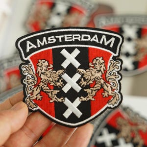 May include: A red, black and white embroidered patch with the text "AMSTERDAM" and a coat of arms featuring two lions and a cross.