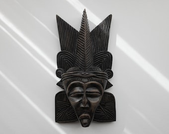 Peut inclure: Un masque mural en bois sculpté à la main de couleur brun foncé, représentant un visage central aux yeux fermés, au petit nez et aux lèvres pleines. Il est surmonté d'une crête pointue en forme de feuille et de motifs géométriques.