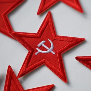 Puede incluir: Parches bordados rojos en forma de estrella con un emblema blanco de martillo y hoz. Varios parches son visibles sobre una superficie blanca. Los parches tienen un aspecto texturizado.