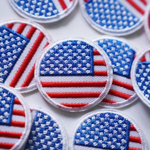 Pode incluir: Uma coleção de pequenos patches redondos bordados com a bandeira americana. Os patches são vermelhos, brancos e azuis com um fundo branco e uma borda vermelha.