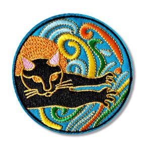 Op de afbeelding: Een ronde geborduurde patch met een zwarte kat die op een blauwe achtergrond ligt met kleurrijke swirls en golven. De kat heeft roze oren en gele ogen.